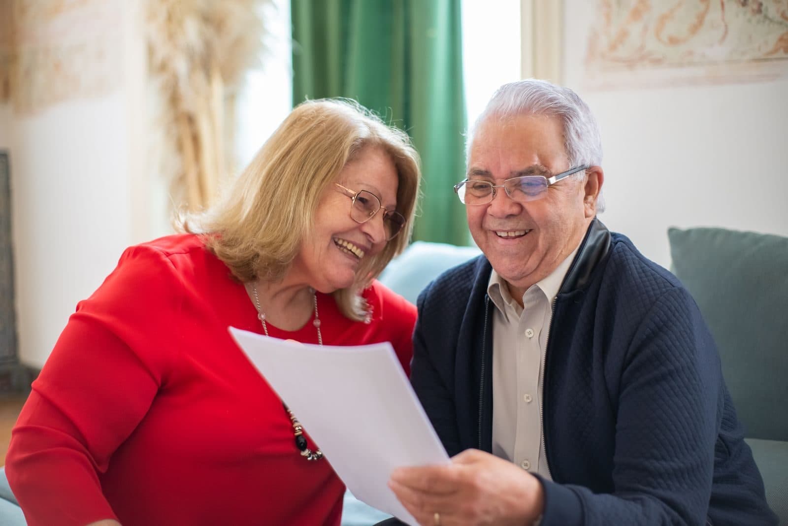 An older couple sitting on a sofa, smiling as they read a document together